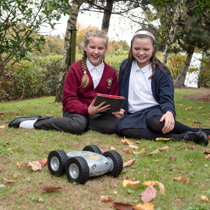 Two young girls in school uniforms sitting on grass with a tablet and a rugged robot toy.